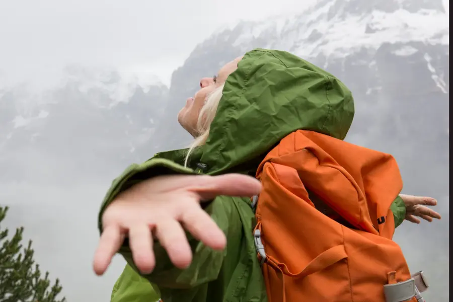 Dame i grønnregnjakke med oransje sekk holder armene ut og ser opp mot himmelen med snødekkede fjell i bakgrunnen
