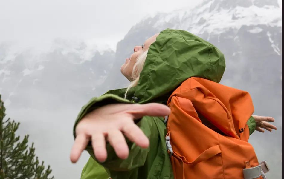 Dame i grønnregnjakke med oransje sekk holder armene ut og ser opp mot himmelen med snødekkede fjell i bakgrunnen