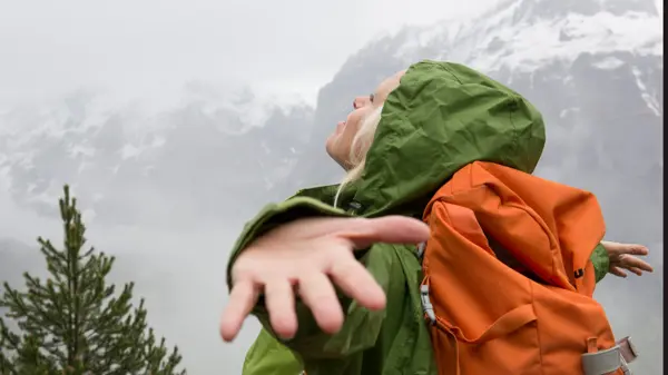Dame i grønnregnjakke med oransje sekk holder armene ut og ser opp mot himmelen med snødekkede fjell i bakgrunnen