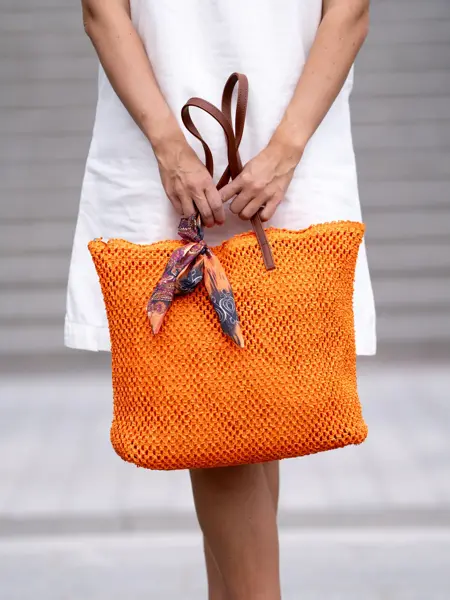 Dame i hvit kjoled holder en orange flettet veske med skinnhanker.