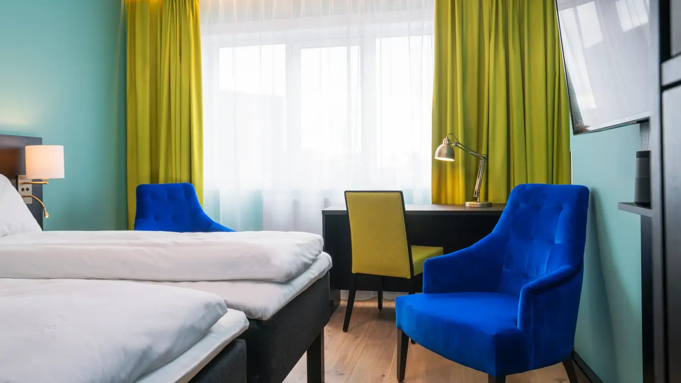 Twin room med hvit sengetøy, gule gardiner, skrivepult og blå lenestol på Thon Hotel Europa i Oslo sentrum rett ved Slottsparken