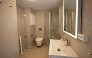 Bildet viser en leilighet i boligprosjektet Otto Ruges vei 80. Badet er i beige toner med toalett, dusjhørne og speil med lys. Bildet er innredet med møbler ved hjelp av KI.