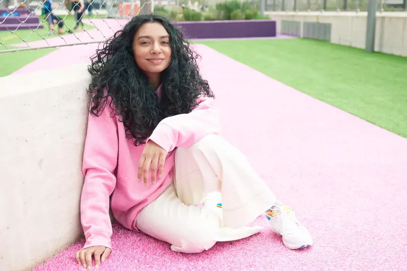 Dame med mørke krøller, rosa hettegenser og lys bukse sitter på rosa asfalt med gress i bakgrunnen 