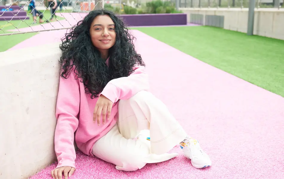 Dame med mørke krøller, rosa hettegenser og lys bukse sitter på rosa asfalt med gress i bakgrunnen 