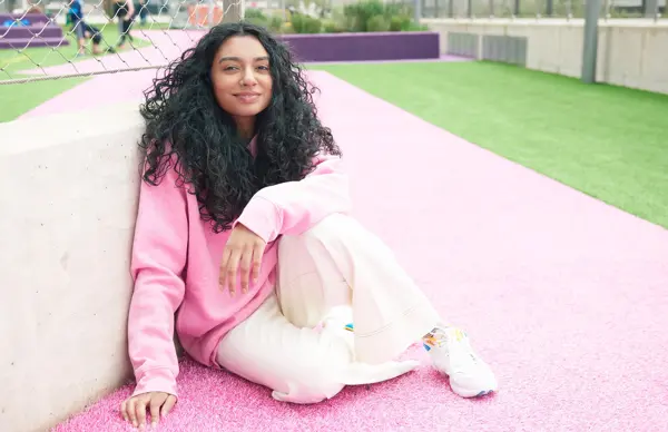 Dame med mørke krøller, rosa hettegenser og lys bukse sitter på rosa asfalt med gress i bakgrunnen 