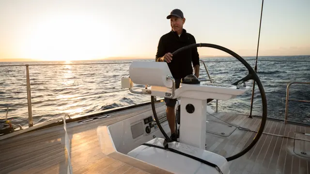 Mann med caps og mørk genser ved roret på en seilbåt på havet i solnedgangen