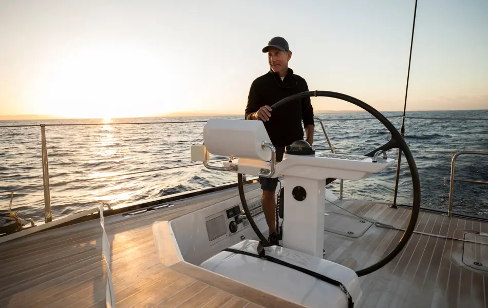 Mann med caps og mørk genser ved roret på en seilbåt på havet i solnedgangen