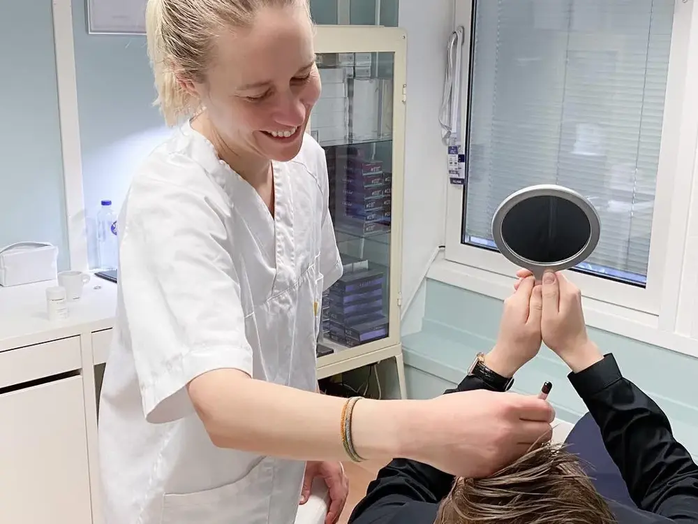 Dame ser seg i håndholdt speil mens smilende pleier holder instrument i pannen 