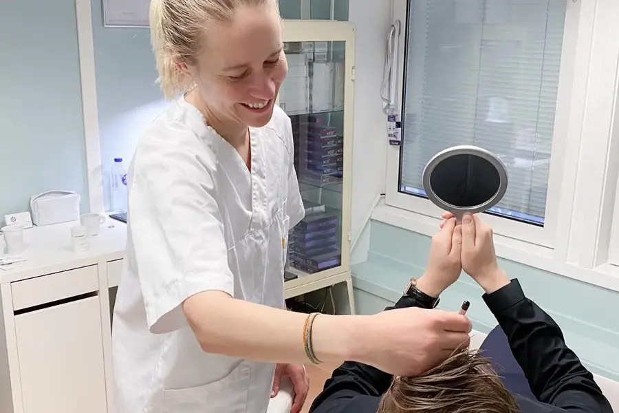 Dame ser seg i håndholdt speil mens smilende pleier holder instrument i pannen 