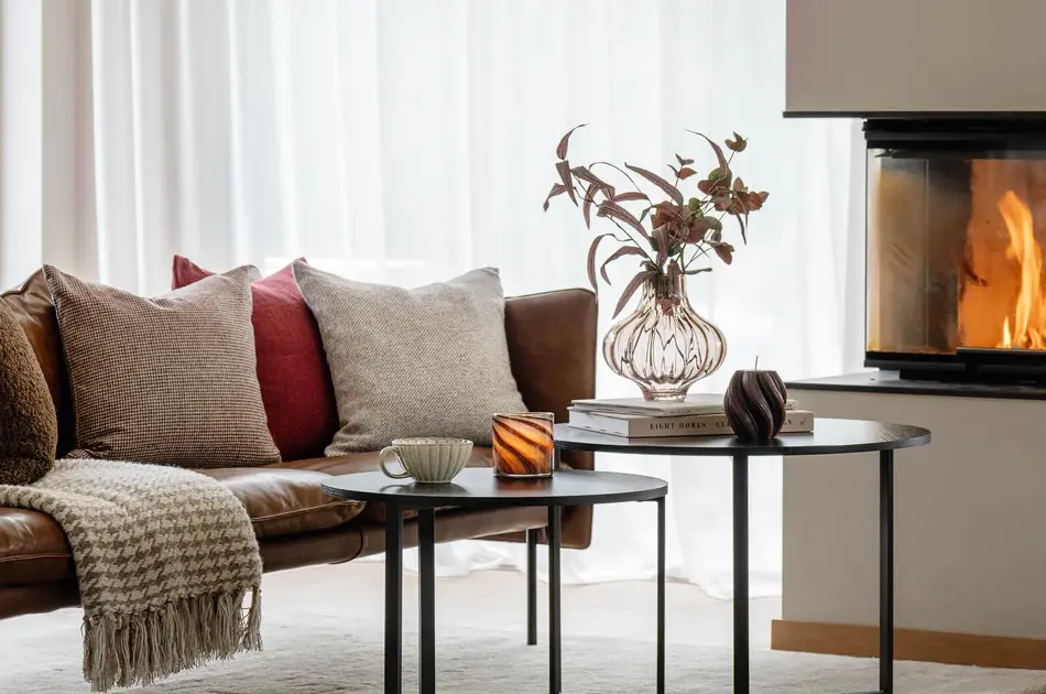 Brun skinnsofa på hvitt teppe med puter og pledd, to sorte sofabord med tekopp, telysholder, vase med blomster og bøker. Peis i bakgrunnen