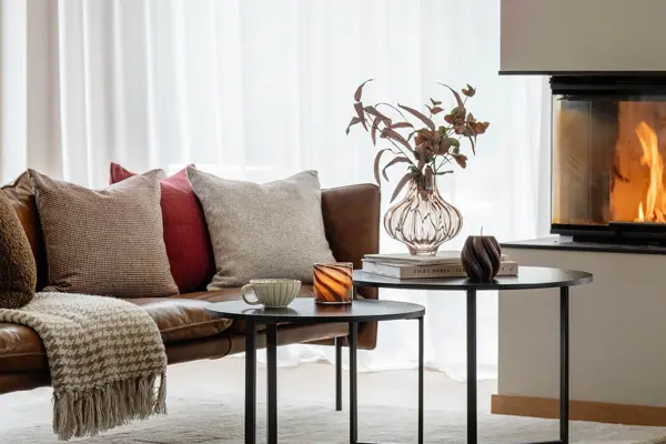 Brun skinnsofa på hvitt teppe med puter og pledd, to sorte sofabord med tekopp, telysholder, vase med blomster og bøker. Peis i bakgrunnen