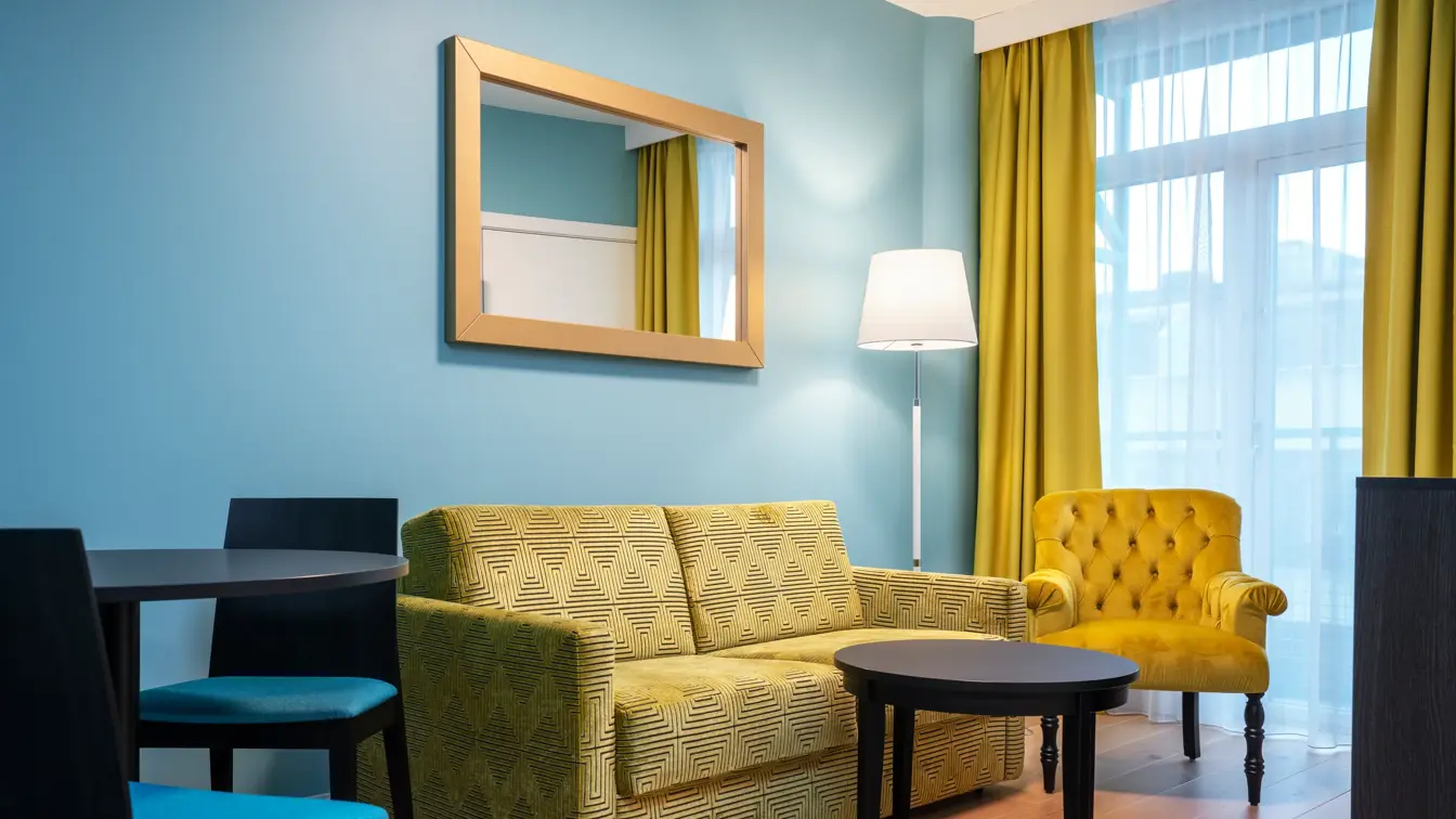 Blå vegger, gule gardiner, vindu fra gulv til tak, spise bord med to stoler, salongbord, gul stol. 