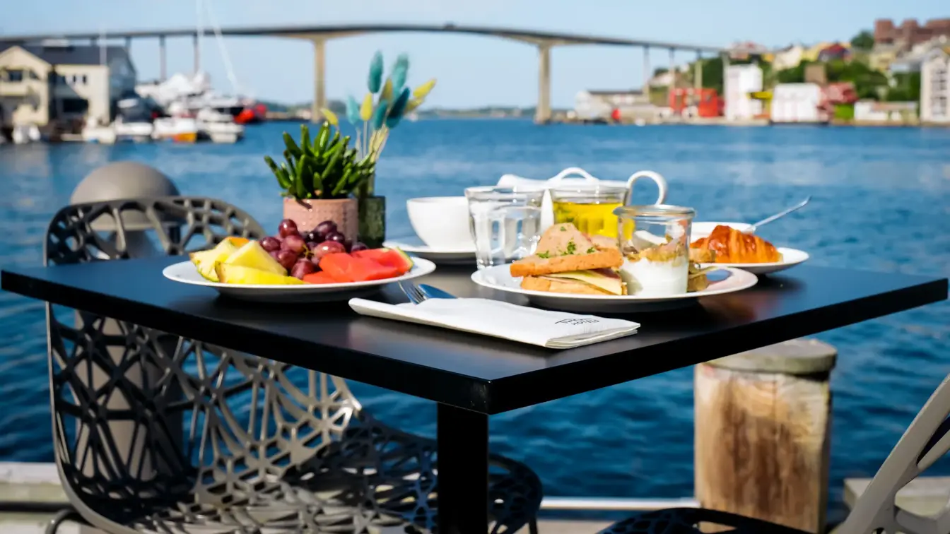 Svart spisebord, frukt, brødmat, croissant, kaffe, juice, syltetøy, frokostbord plassert ute. Havet i bakgrunn