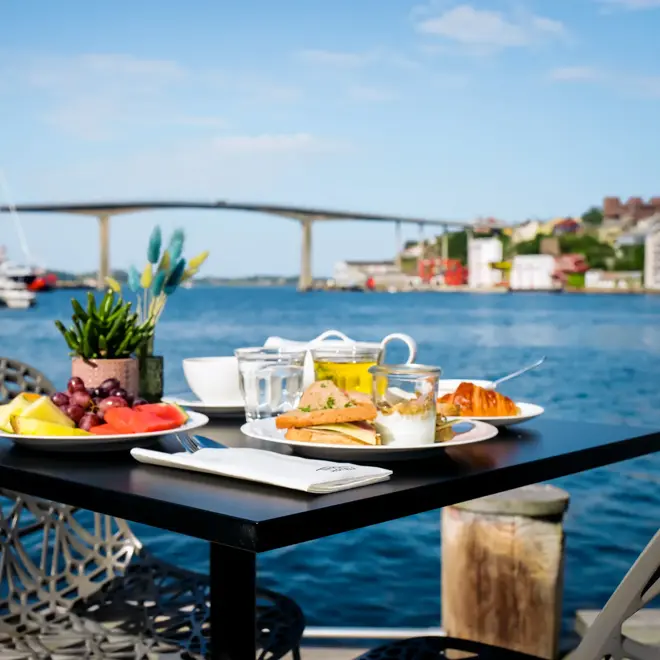 Svart spisebord, frukt, brødmat, croissant, kaffe, juice, syltetøy, frokostbord plassert ute. Havet i bakgrunn