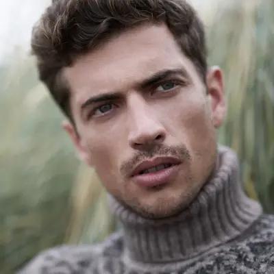 Mann i høyhalset strikkegenser sitter i gresset og ser bort