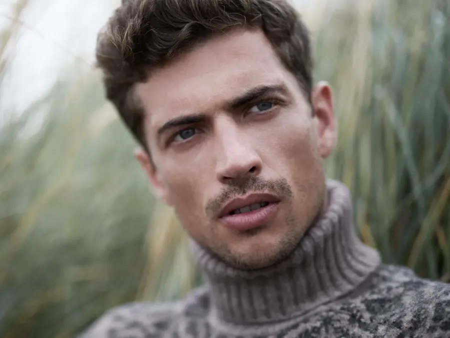 Mann i høyhalset strikkegenser sitter i gresset og ser bort