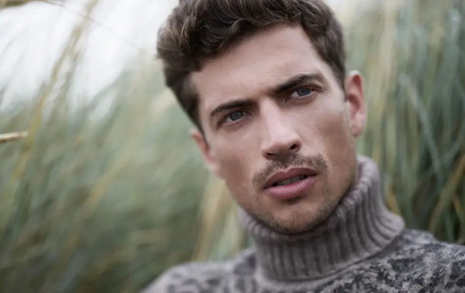 Mann i høyhalset strikkegenser sitter i gresset og ser bort