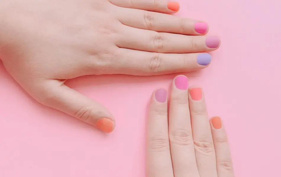 Rosa bakgrunn og to hender med neglelakk i ulike pastellfarger, lilla, rosa og oransje. 