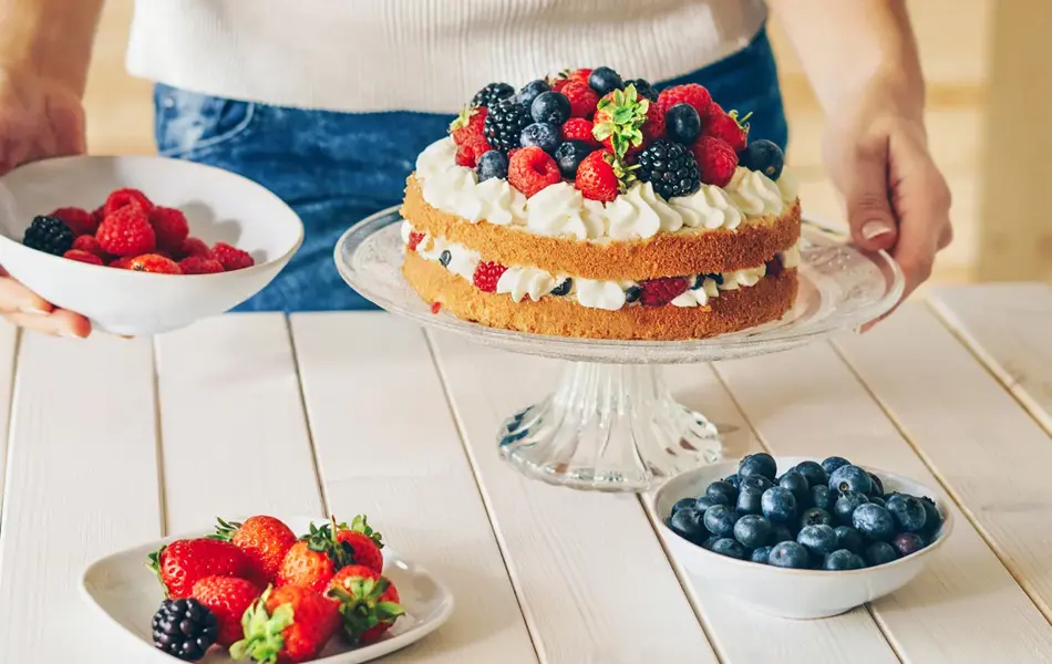 Nærbilde av hender som presenterer kake med krem og skogsbær. Foto til artikkel om dekorasjoner til 17. mai.