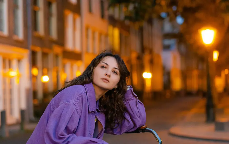 Dame med mørk lilla olajakke og sort bukse sitter på sykkel med hus og trær i bakgrunnen