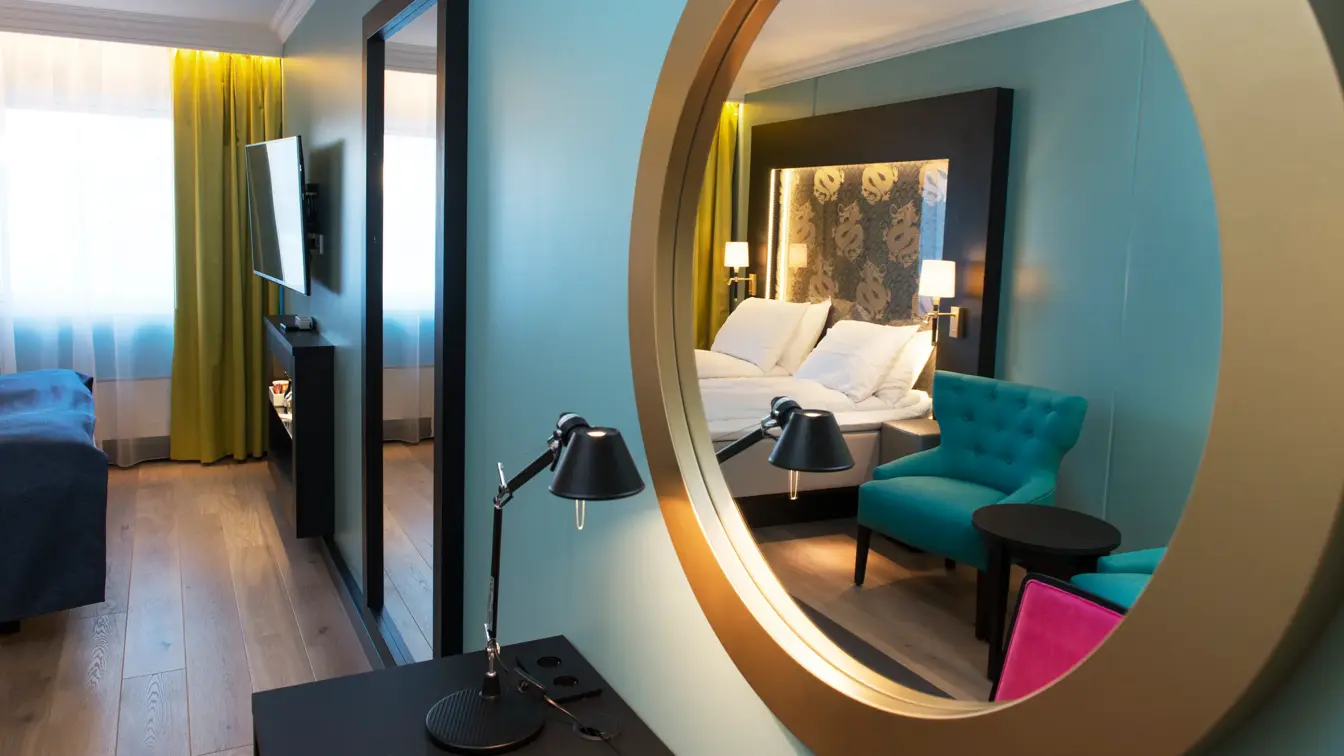 Rundt speil med speilbilde av dobbeltseng og blå lenestoler på Thon hotel Terminus i Oslo sentrum nær Jernbanetorget