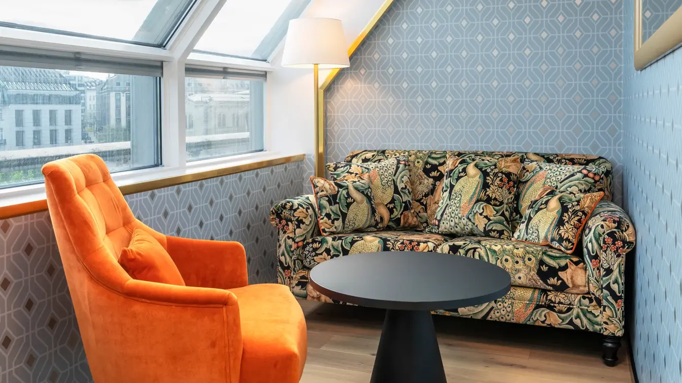 Sittegruppe med fargerik sofa, oransje stol, sort rundt bord, mønstre sølvtapet og store vinduer med utsikt i junior suite på Thon Hotel Cecil