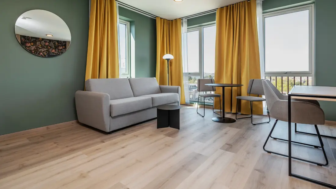 Leilighet med grå sofa, spiseplass, skrivebord med tilhørende stol, parkett, grønne vegger, store vinduer og gule gardiner.