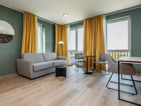 Leilighet med grå sofa, spiseplass, skrivebord med tilhørende stol, parkett, grønne vegger, store vinduer og gule gardiner.
