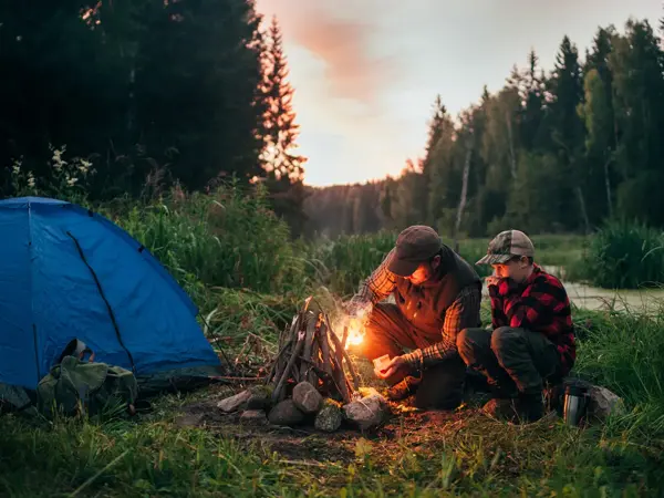Mann og gutt sitter ved bål i kveldssol ved siden av teltet. Foto til artikkel om gaver til pappatyper på farsdag. 
