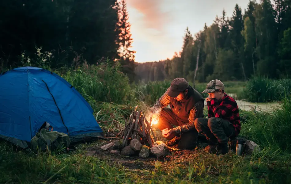 Mann og gutt sitter ved bål i kveldssol ved siden av teltet. Foto til artikkel om gaver til pappatyper på farsdag. 