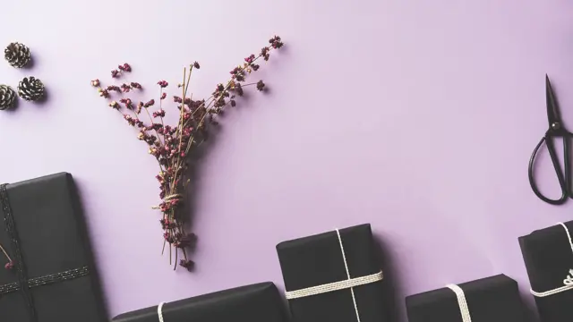 Black wrapped Christmas gift box flat lay with flower branches, scissors on purple background.
