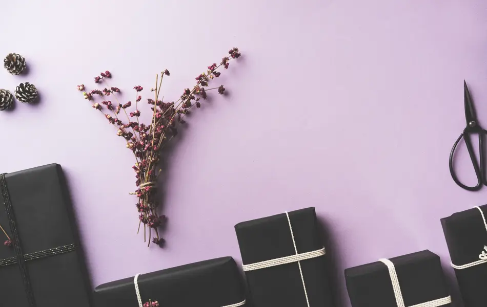 Black wrapped Christmas gift box flat lay with flower branches, scissors on purple background.