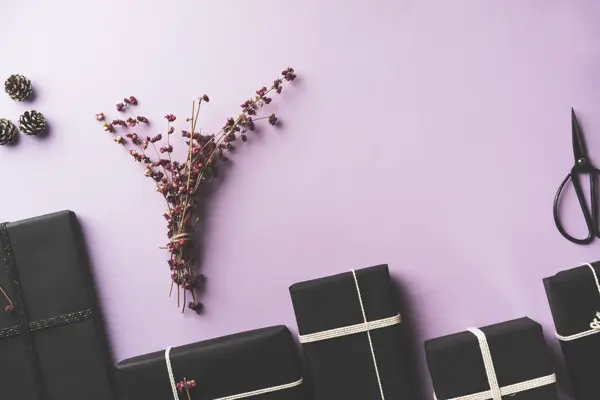Black wrapped Christmas gift box flat lay with flower branches, scissors on purple background.