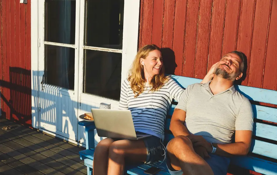 Mann med grå t-skjorte med v-hals sitter sammen med dame på blå benk i hytteveggen