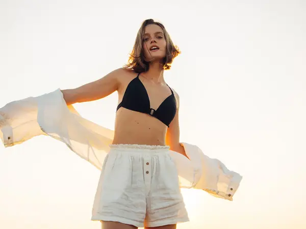 Dame med sort bikini og hvit linshorts og skjorte poserer foran kamera  