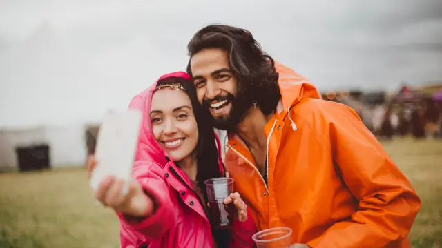 Young couple are at a music festival in rainy weather. They are wearing colourful rain macs and are using a smartphone to take a selfie of them together.
