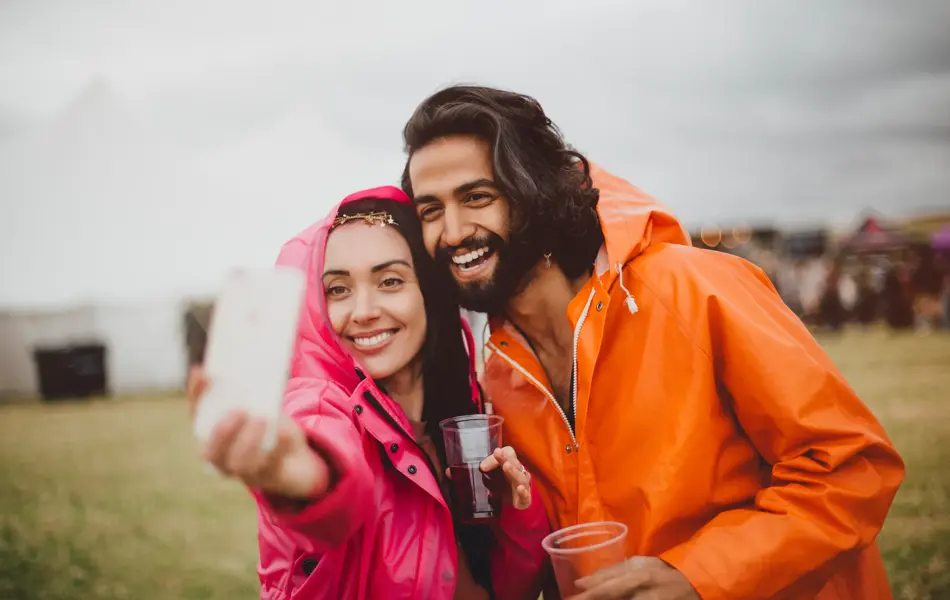 Young couple are at a music festival in rainy weather. They are wearing colourful rain macs and are using a smartphone to take a selfie of them together.