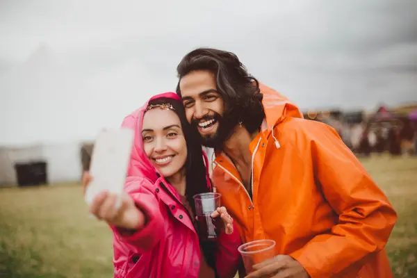 Young couple are at a music festival in rainy weather. They are wearing colourful rain macs and are using a smartphone to take a selfie of them together.