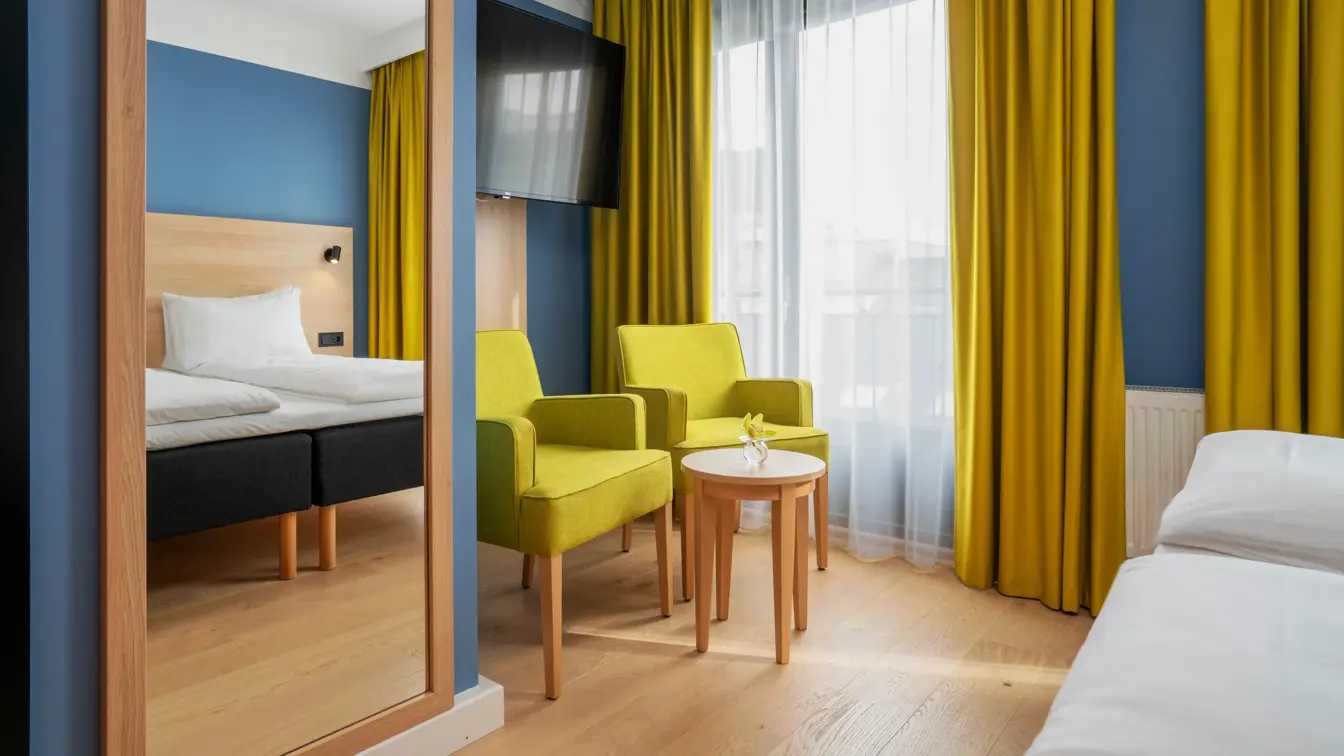 Speil, sittegruppe med to gule stoler, bord og TV, blå vegger og gule gardiner på dobbeltrom på Thon Hotel Astoria. 