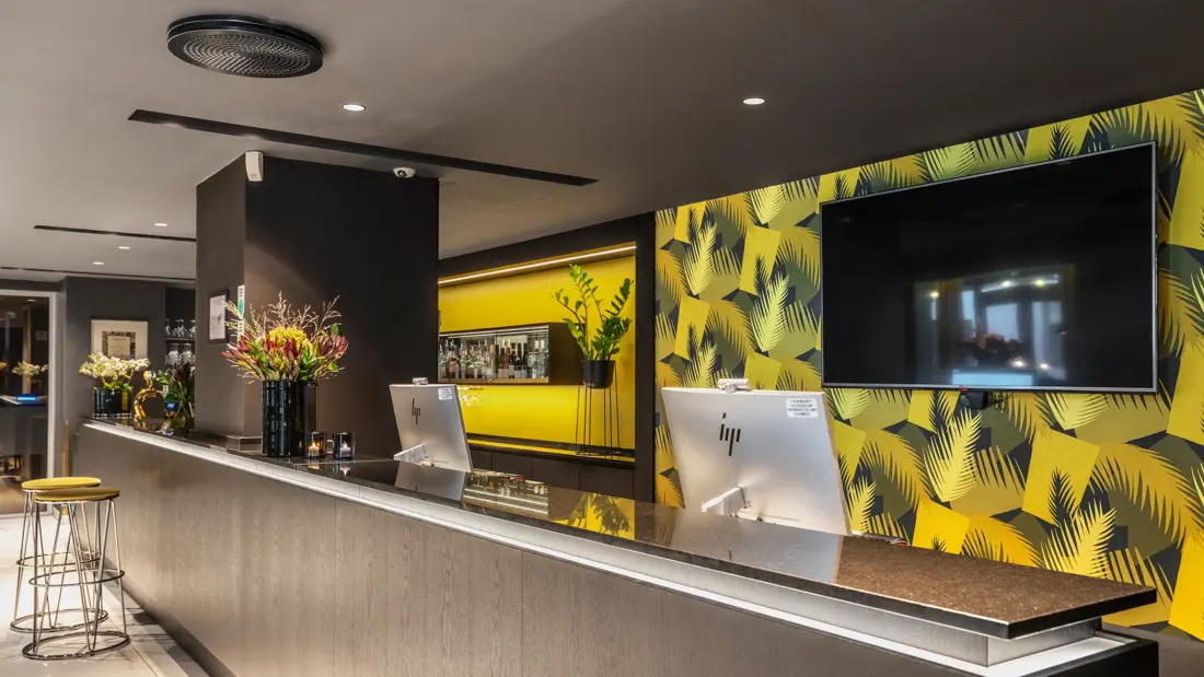 Resepsjonsdisk med to PC skjermer, gul mønstrete tapet og TV skjerm.