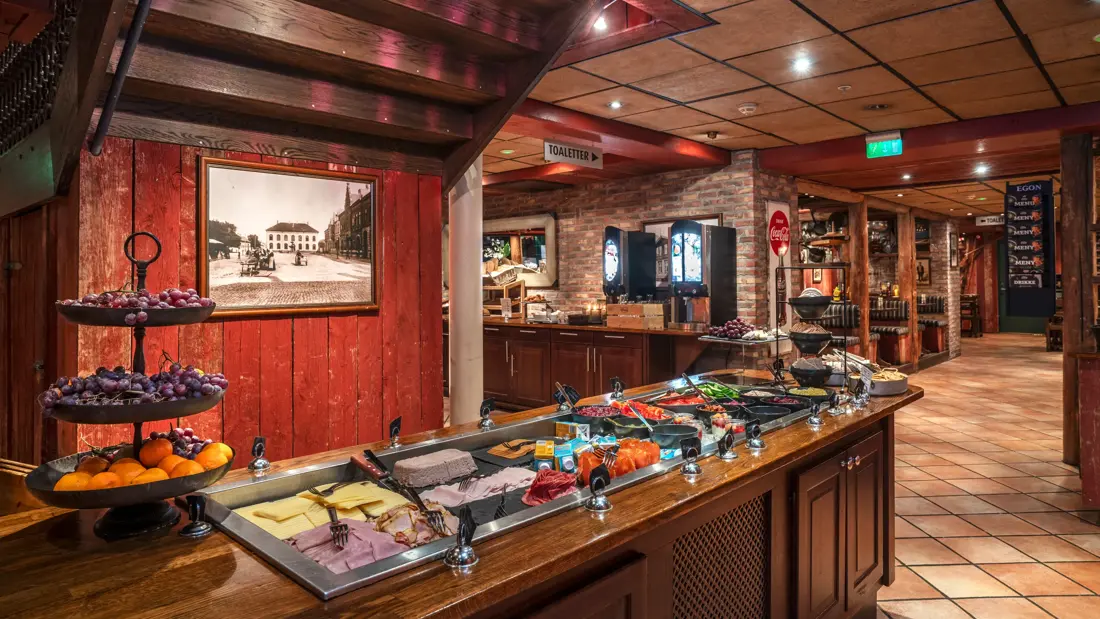 Frokostbuffé med frukt, ost og kjøttpålegg. I bakgrunnen ses dispensere med ulik drikke, brød og sitteplasser.