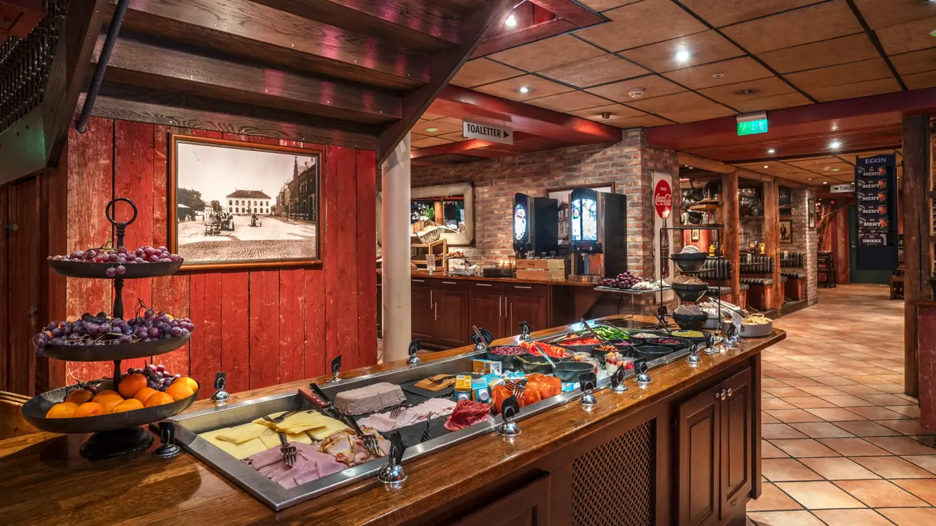 Frokostbuffé med frukt, ost og kjøttpålegg. I bakgrunnen ses dispensere med ulik drikke, brød og sitteplasser.