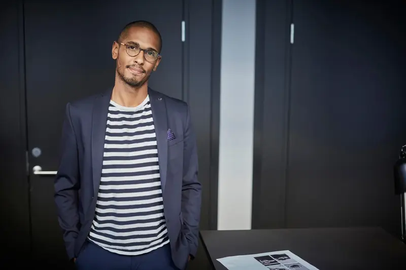 Mann med blå dress, hvitstripete t-skjorte og briller poserer til kamera inne på kontor 