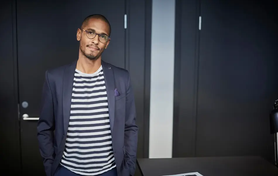 Mann med blå dress, hvitstripete t-skjorte og briller poserer til kamera inne på kontor 