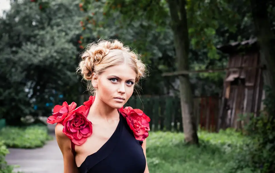 Kvinnelig modell står utendørs i en grønn hage og holder på et gjerde. Modellen har oppsatt hår og er iført en kreasjon med roser og en svart drakt.