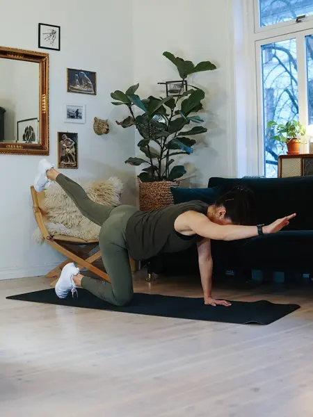treningsekspert Anna Johansen trener på matte på gulvet, mørkegrønn sofa i bakgrunnen, lys stol med pels oppi, str grønn plante, bilder på veggen