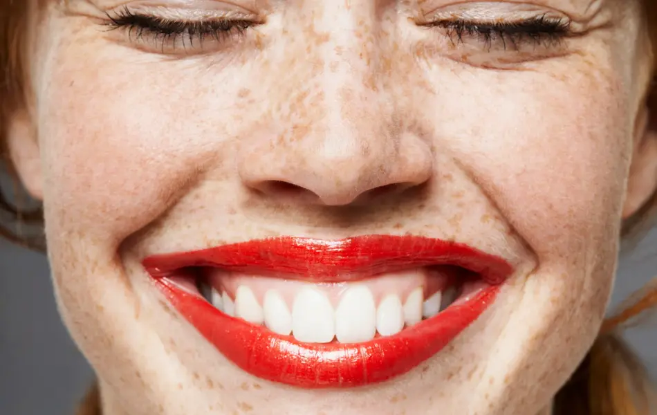 Nærbilde av dame med pannelugg og knallrøde lepper som smiler med lukkede øyne