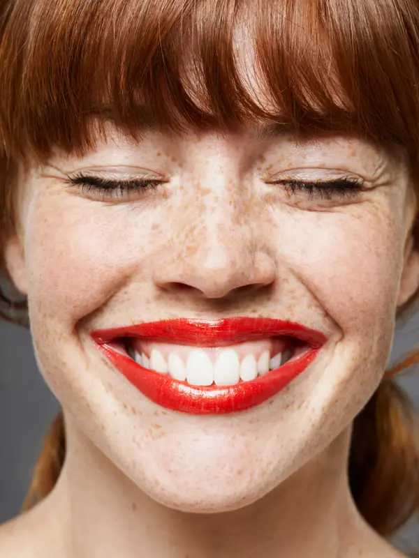 Nærbilde av dame med pannelugg og knallrøde lepper som smiler med lukkede øyne