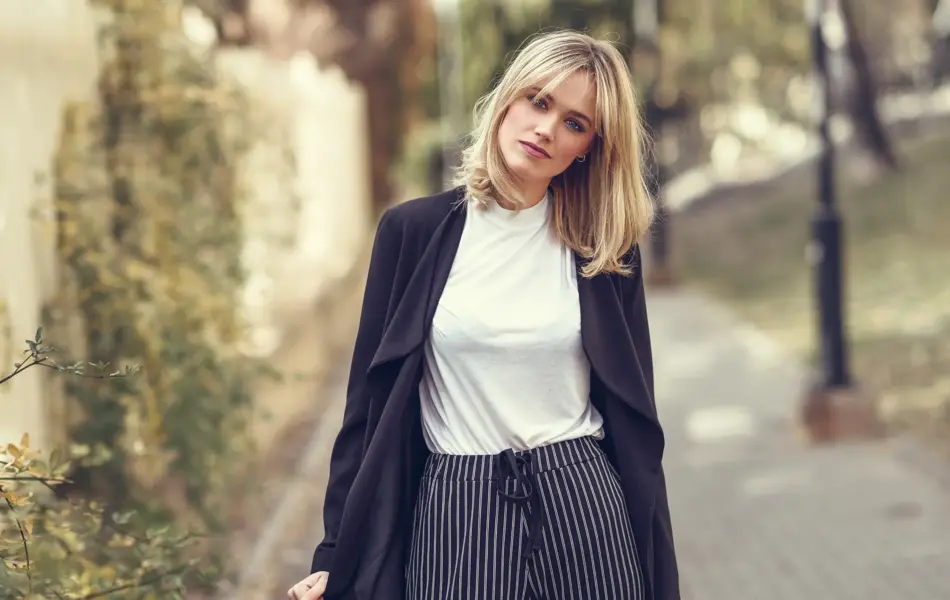 Beautiful blonde woman in urban background. Young girl wearing black blazer jacket and striped trousers standing in the street. Pretty female with straight hair hairstyle and blue eyes.