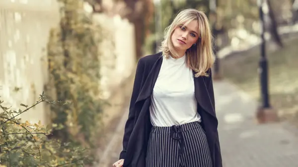 Beautiful blonde woman in urban background. Young girl wearing black blazer jacket and striped trousers standing in the street. Pretty female with straight hair hairstyle and blue eyes.